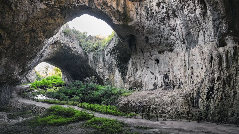 A natural rock arch formation with a dirt path and greenery in the foreground.
