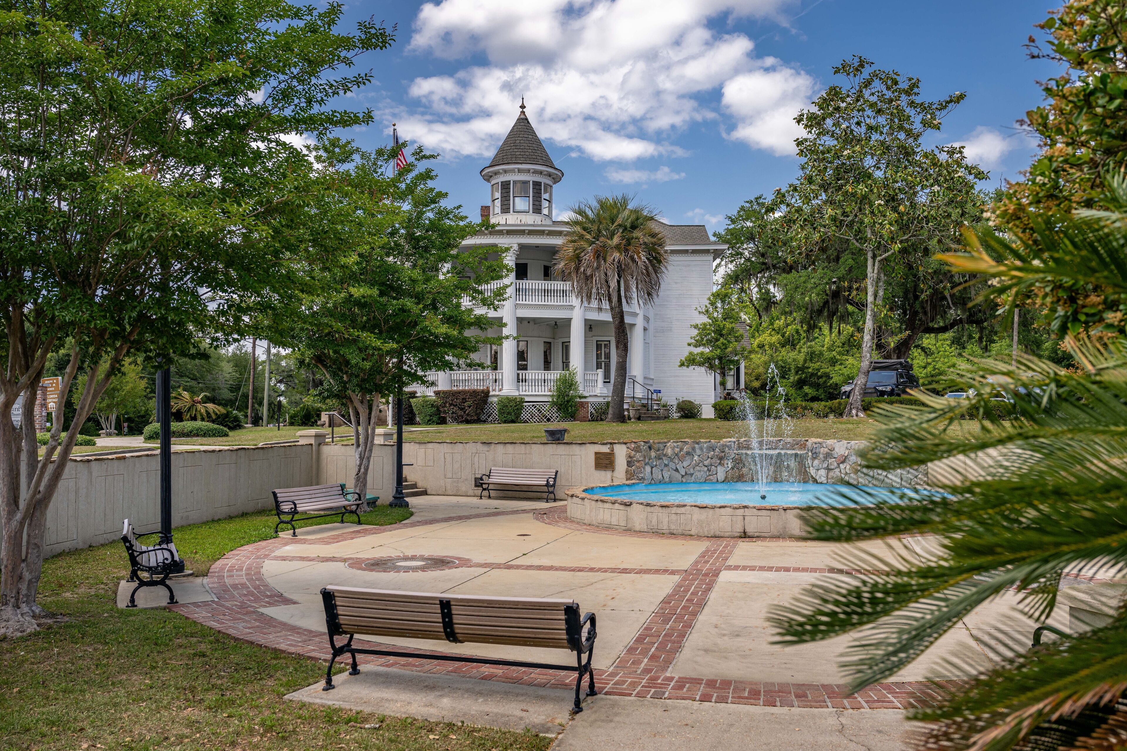 A large white building with a tower, surrounded by a park with benches, a fountain, and palm trees.