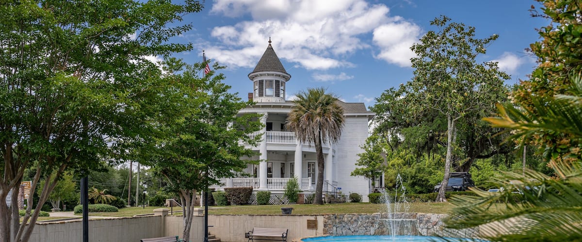 A large white building with a tower, surrounded by a park with benches, a fountain, and palm trees.