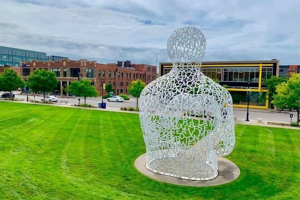 En stor, hvid skulptur af et menneske i en park med bygninger i baggrunden.