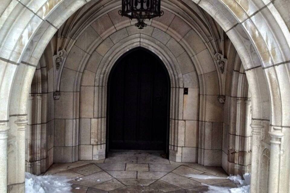 An arched stone doorway with a black door, surrounded by hexagonal tiles and snow on the ground.