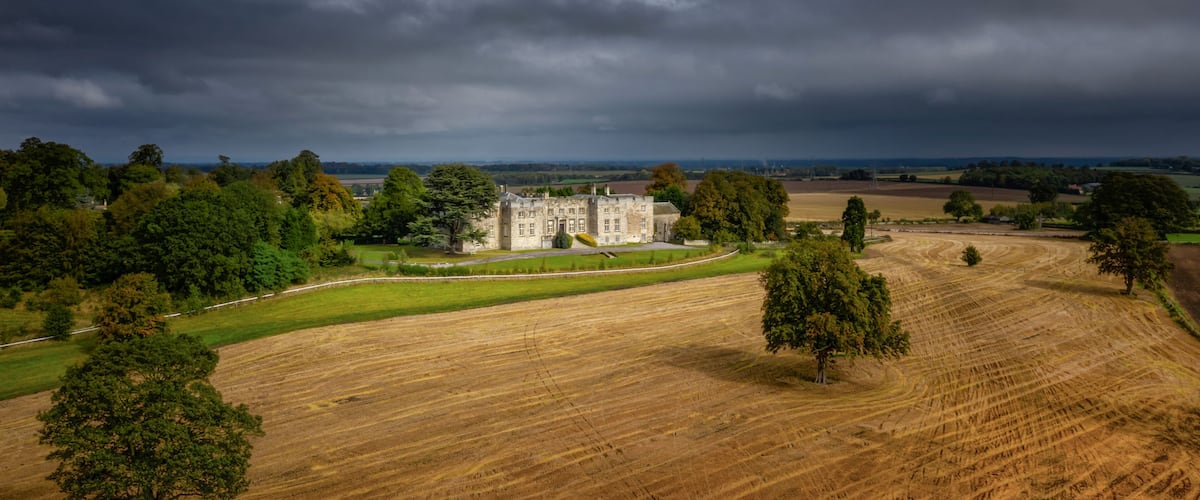 A large estate with a prominent stone building, expansive fields, and a dramatic cloudy sky.