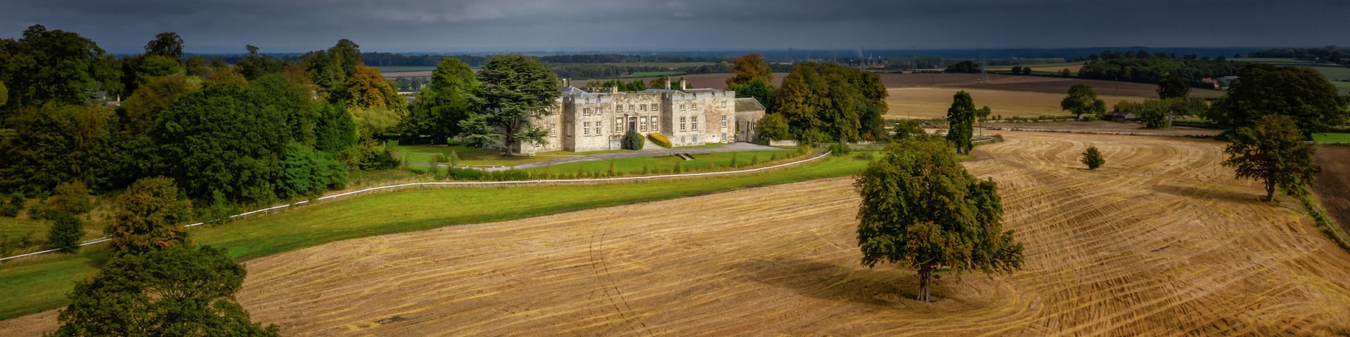 A large estate with a prominent stone building, expansive fields, and a dramatic cloudy sky.