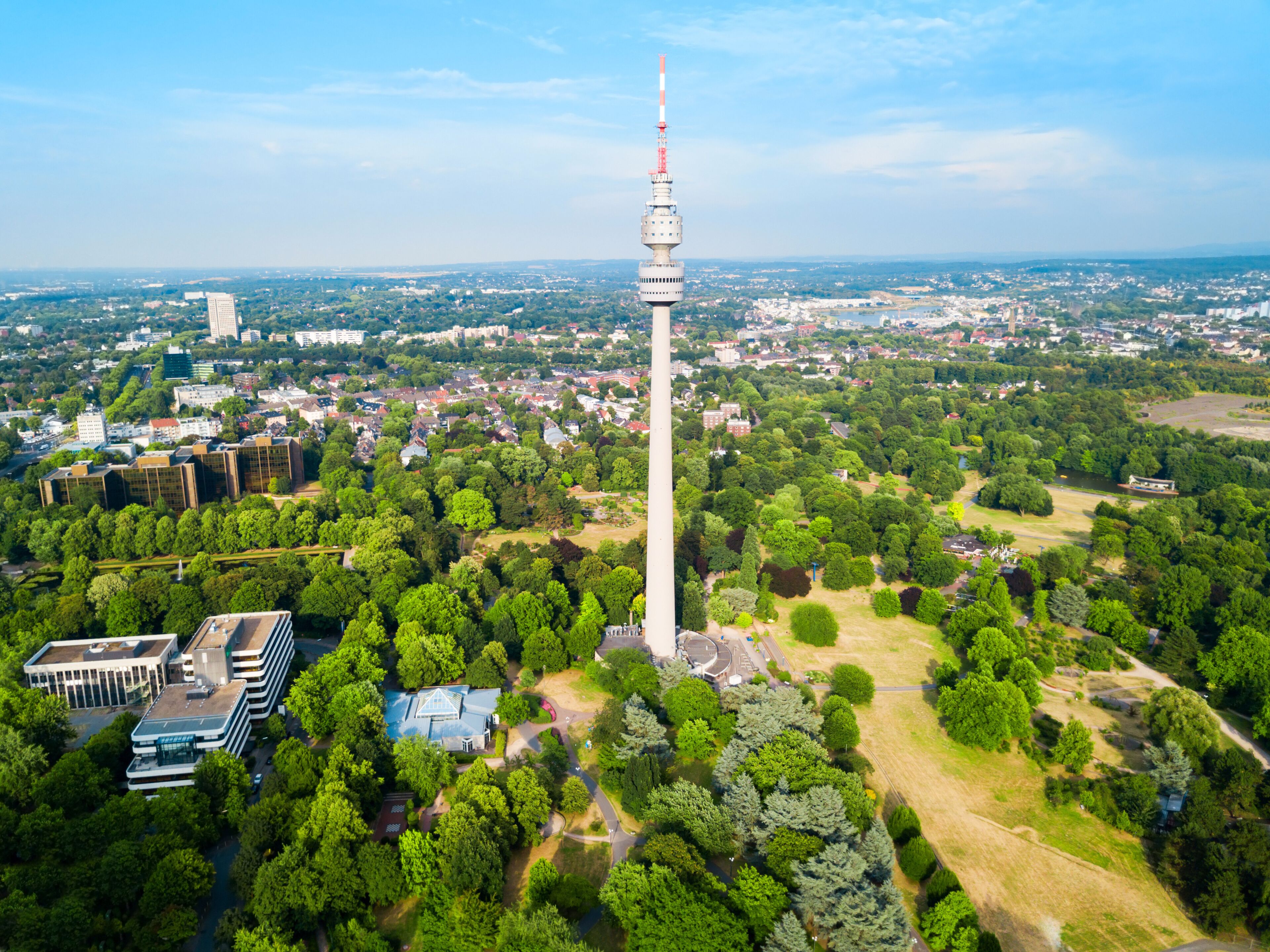 A tower with a red top, surrounded by a dense forest and a cityscape in the background.