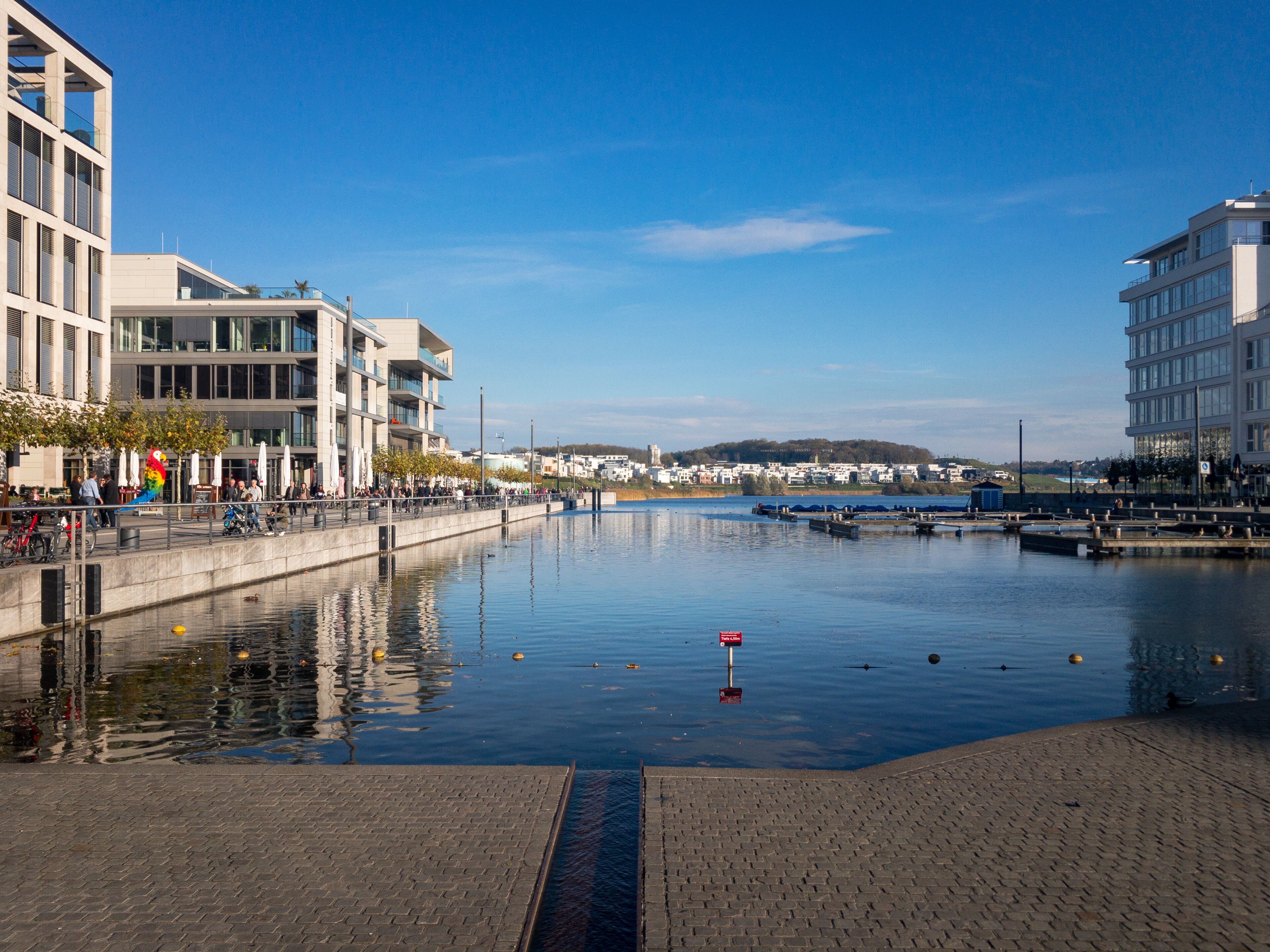 Un paseo marítimo con edificios modernos, un río tranquilo y un cielo azul despejado.