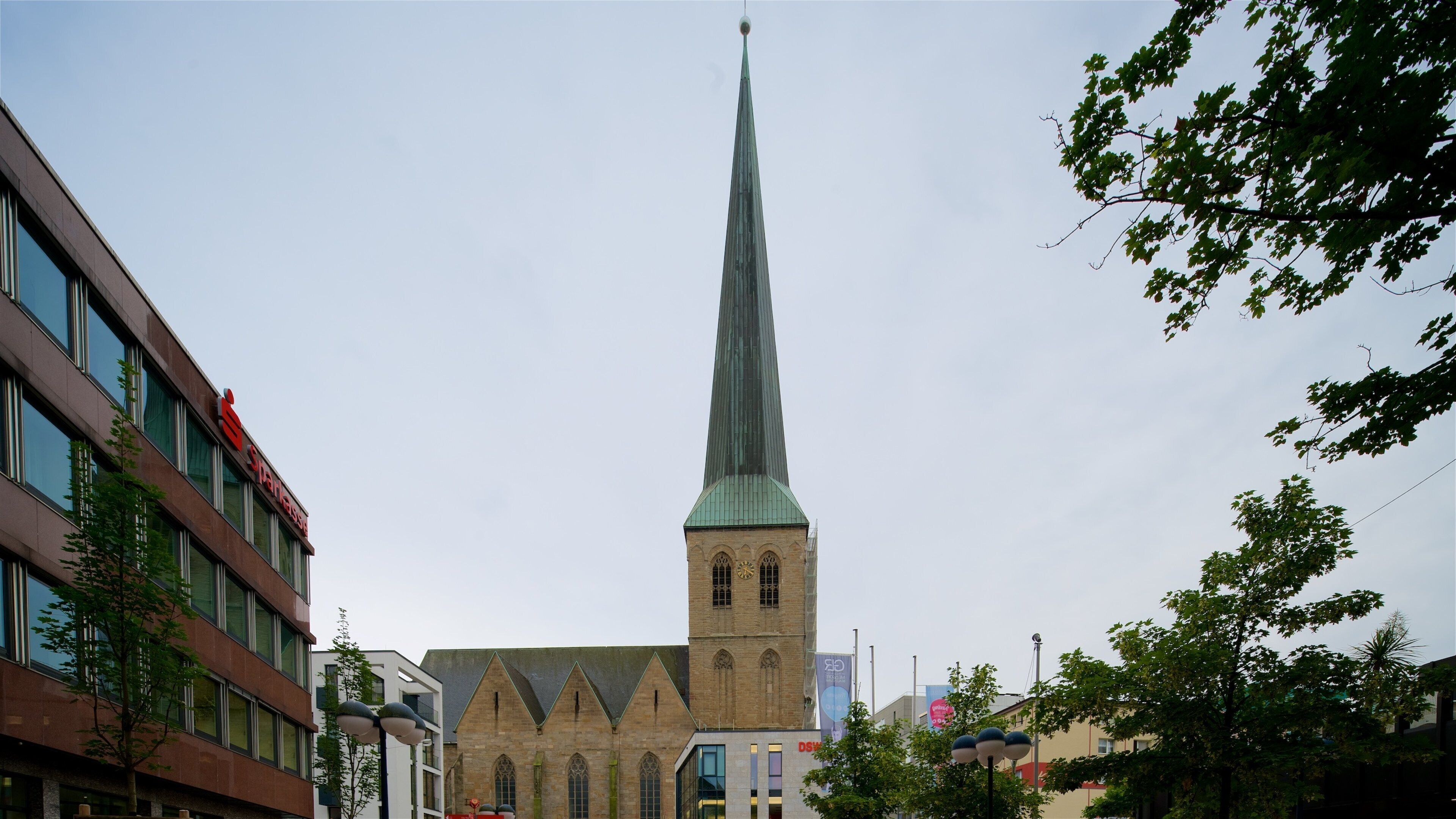 A church with a tall spire, a modern building with a red sign, and trees in the foreground.