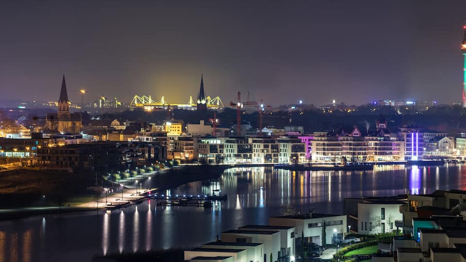 A nighttime cityscape with illuminated buildings, a river, and a bridge.