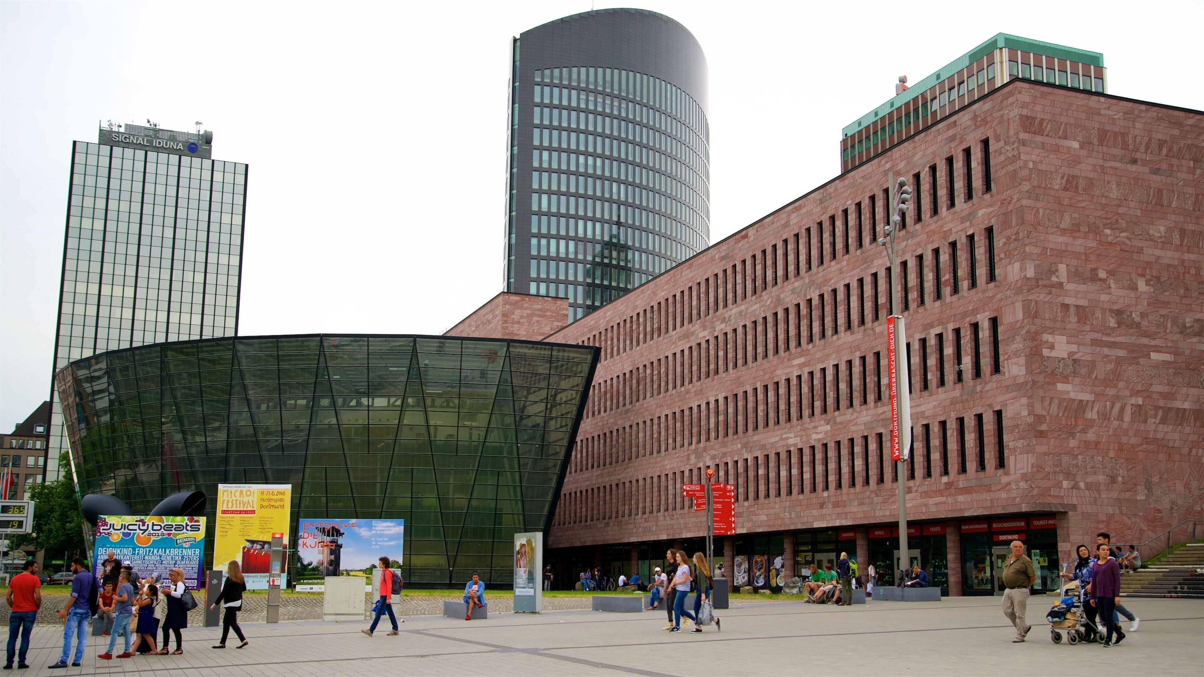A modern urban square with a curved glass building, a red brick building, and tall skyscrapers in the background.