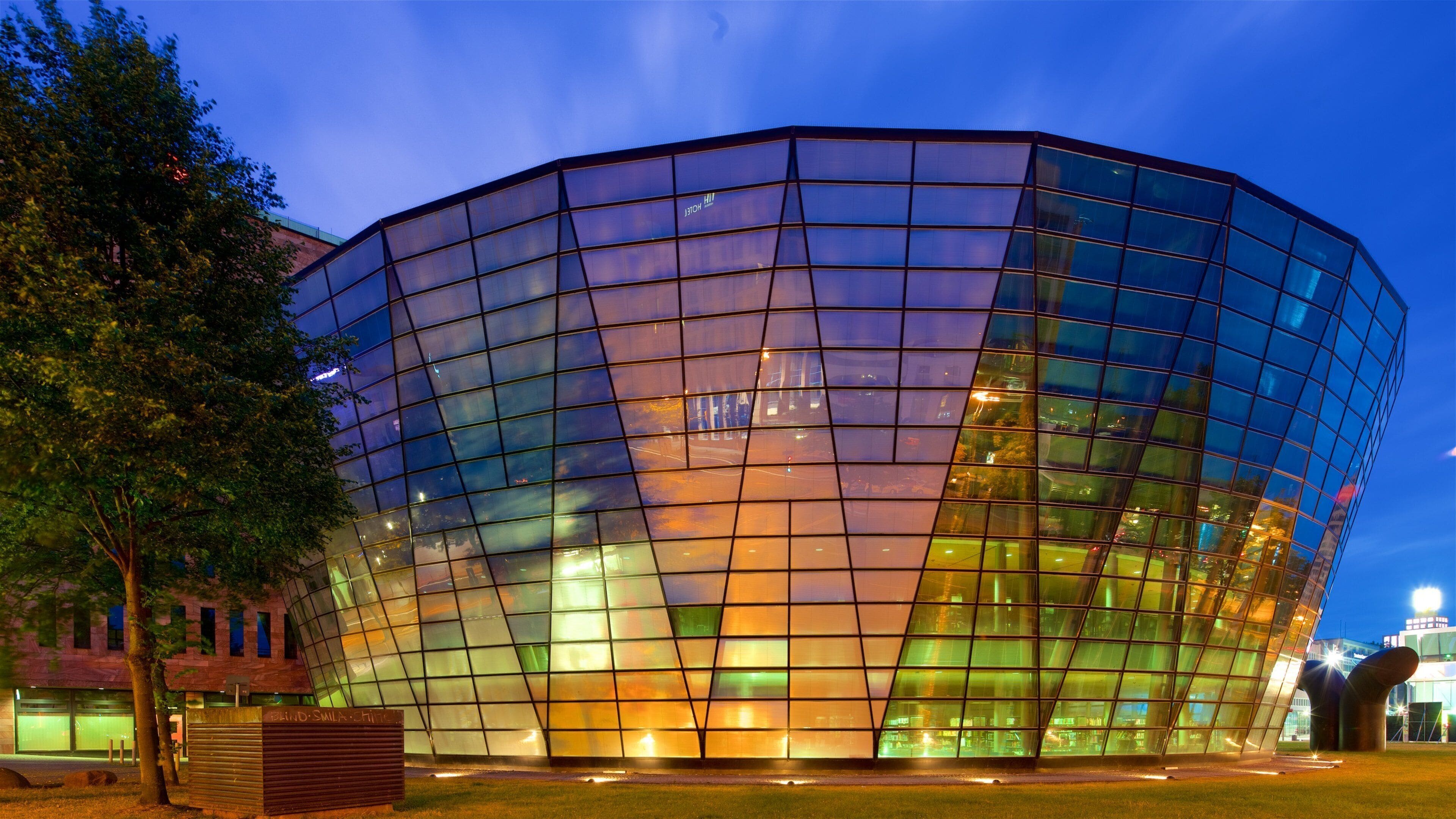 A modern glass building with a curved facade, surrounded by trees and a grassy area.