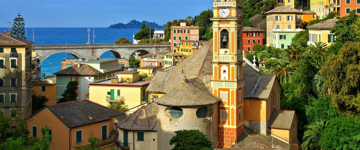 A coastal town with colorful buildings, a prominent church tower, and a bridge over the sea.