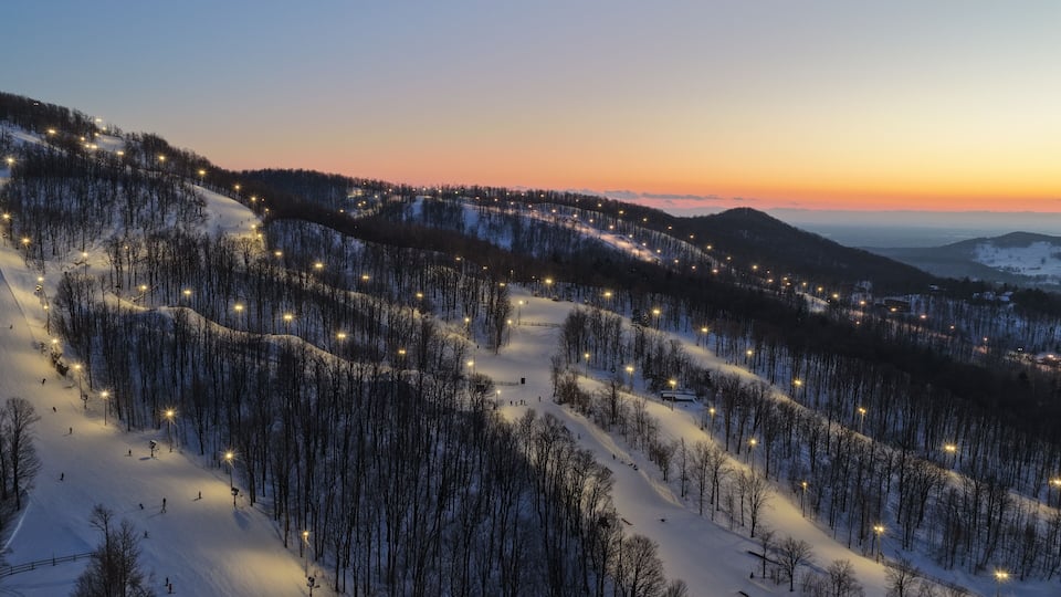 A snow-covered ski slope at dusk with artificial lights illuminating the trees and paths.
