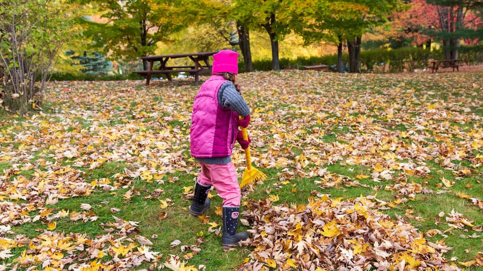 A child in a pink vest and hat collecting leaves in a park with trees and a picnic table.