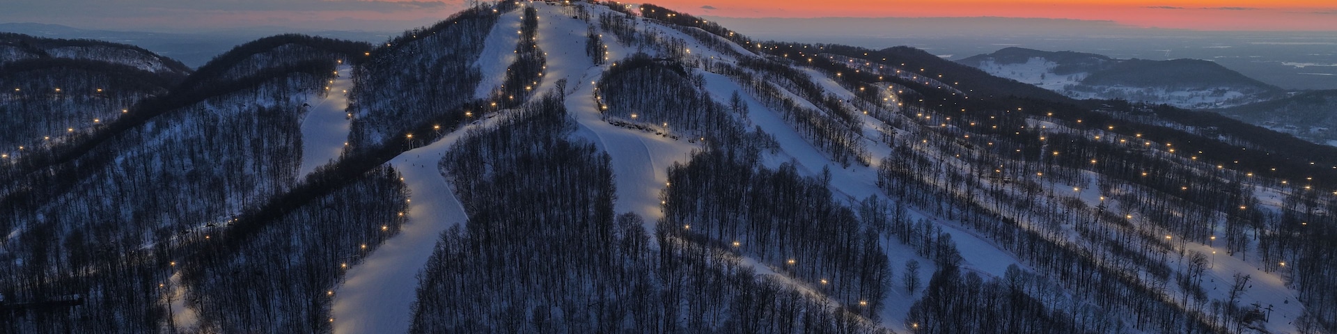 Un paisaje montañoso nevado al atardecer, con pistas de esquí iluminadas y un edificio distante.
