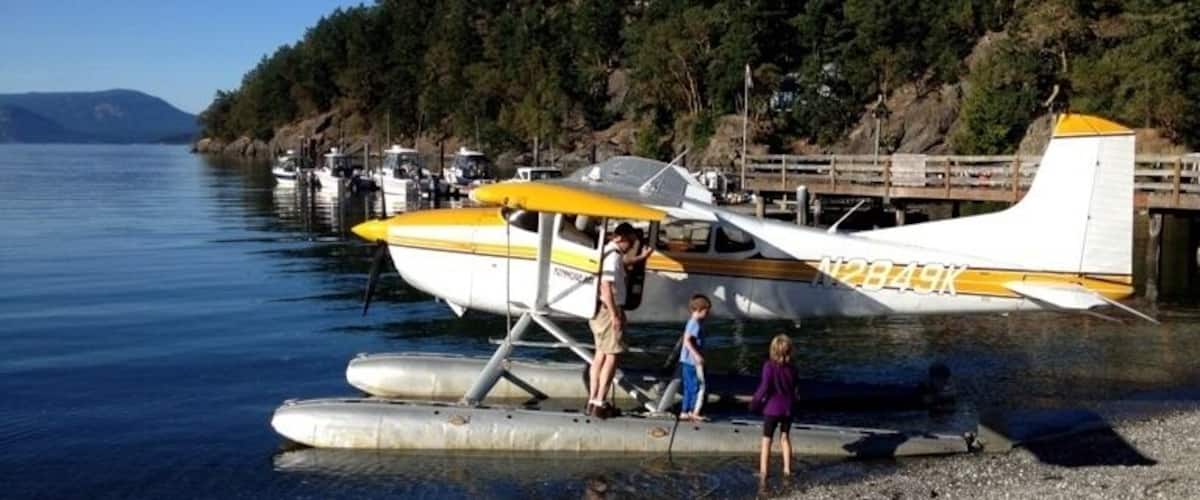 A seaplane parked on a pebbly shore with a forested hill in the background.