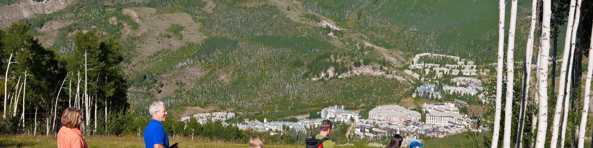 Um grupo de pessoas caminhando numa colina com relva, com vista para uma vila e montanhas ao fundo.