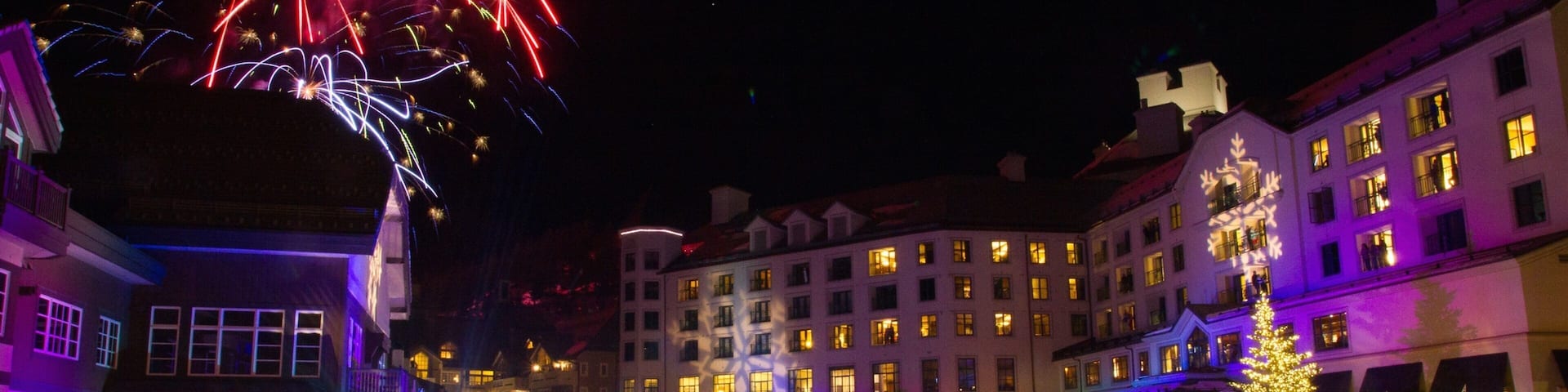 A nighttime outdoor event with fireworks, illuminated buildings, and a crowd gathered around a central stage.