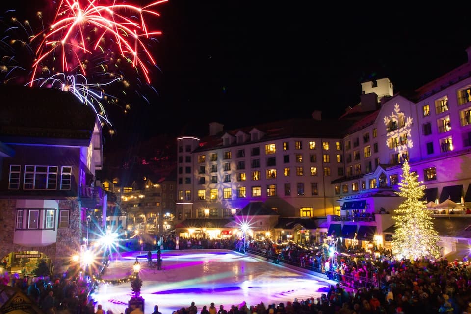 A nighttime outdoor event with fireworks, illuminated buildings, and a crowd gathered around a central stage.