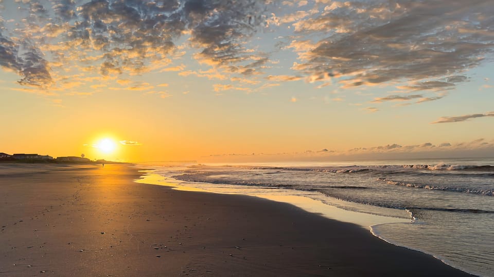A beach at sunset with the sun low on the horizon, waves gently lapping the shore, and a pier in the distance.