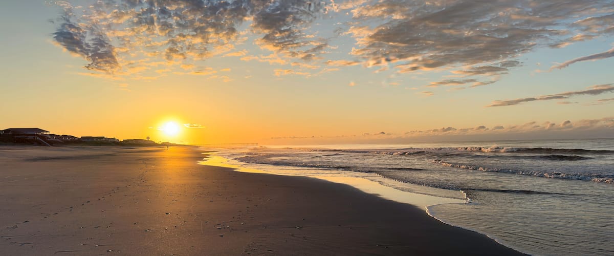A beach at sunset with the sun low on the horizon, waves gently lapping the shore, and a pier in the distance.