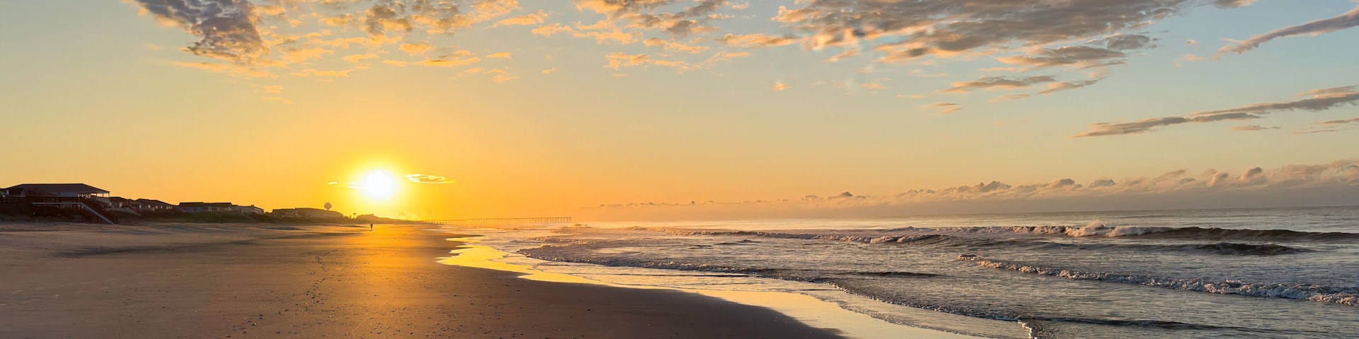 A beach at sunset with the sun low on the horizon, waves gently lapping the shore, and a pier in the distance.