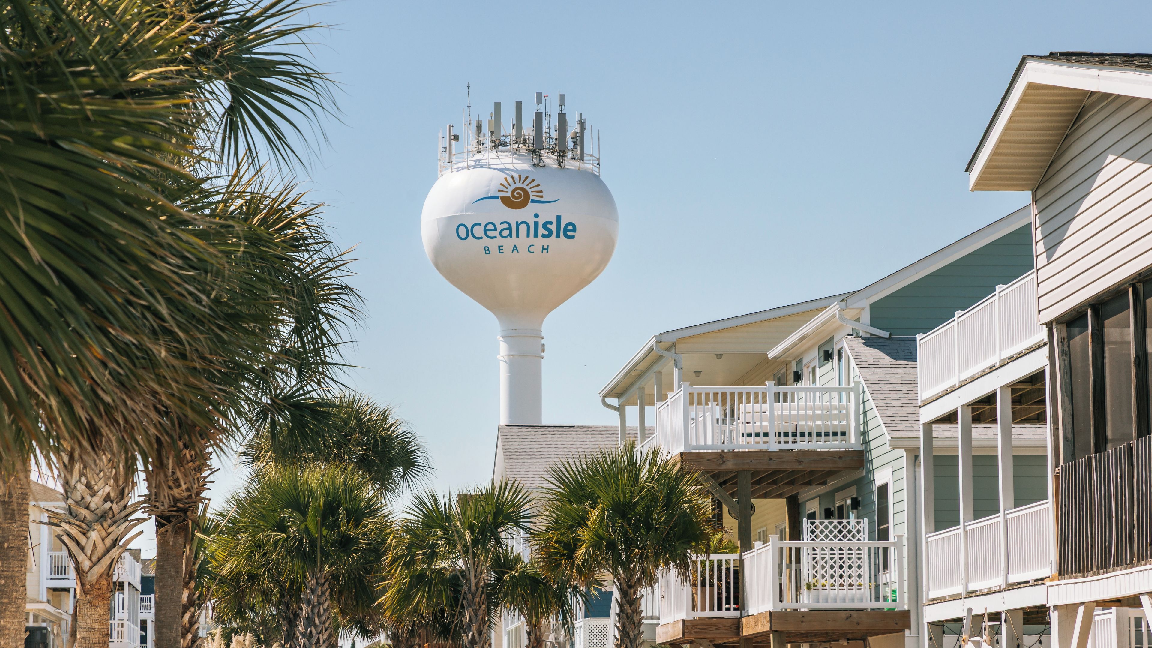A water tower with 'Oceanisle Beach' written on it, surrounded by palm trees and beachfront buildings.