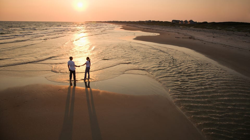 A beach at sunset with two people holding hands, a calm sea, and distant houses.
