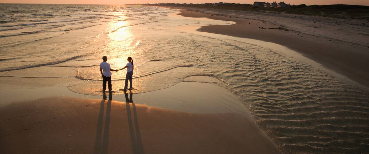 A beach at sunset with two people holding hands, a calm sea, and distant houses.