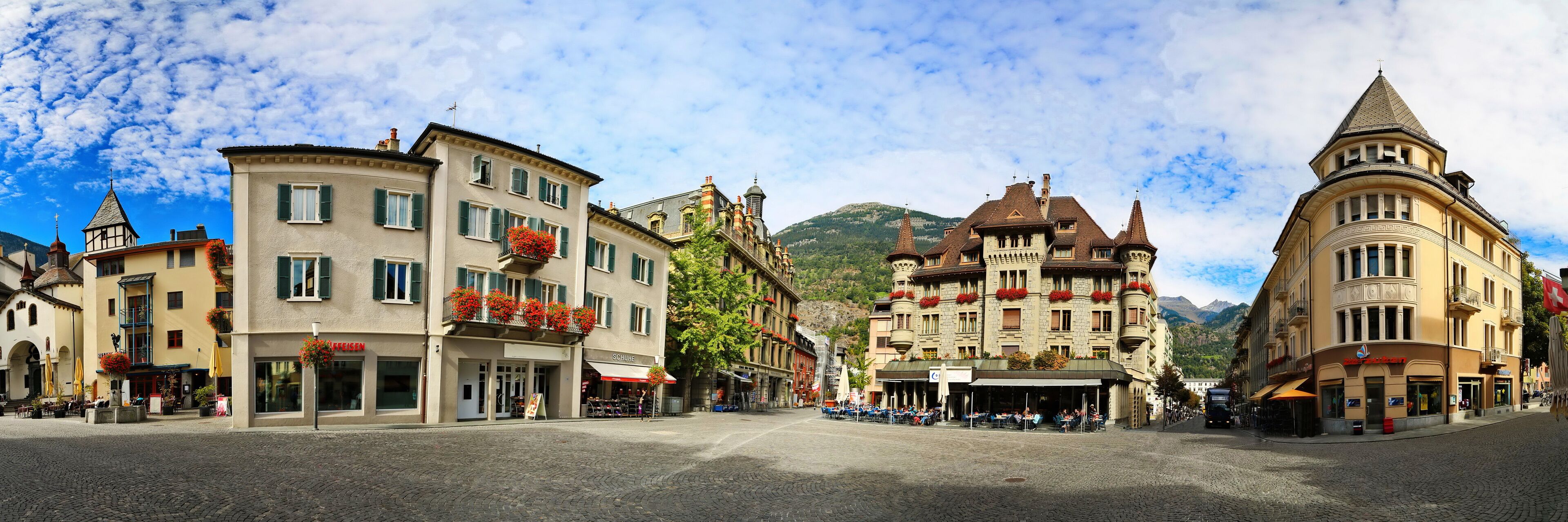 A cobblestone square with historic buildings, a mountain in the background, and outdoor cafes.