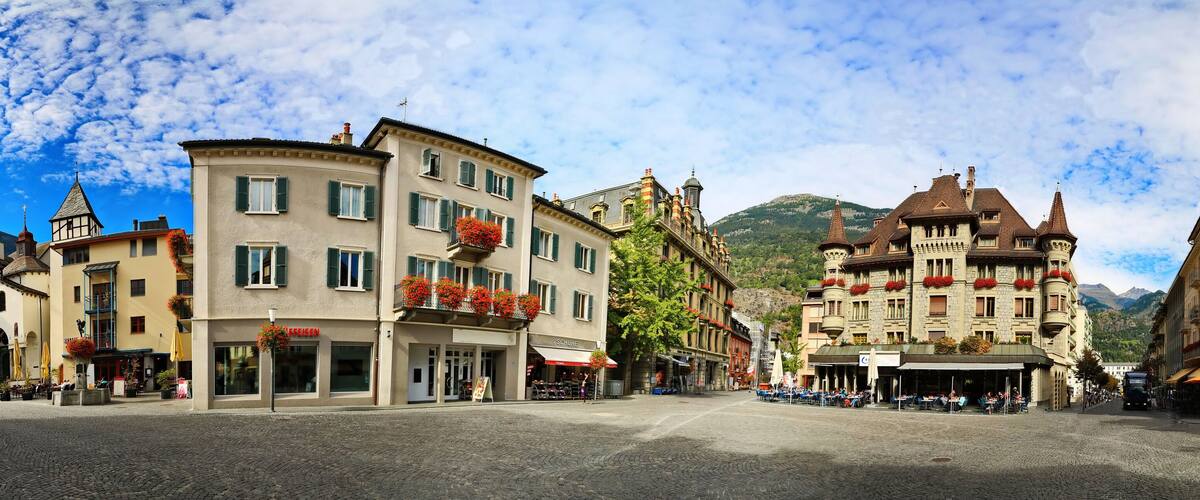 A cobblestone square with historic buildings, a mountain in the background, and outdoor cafes.