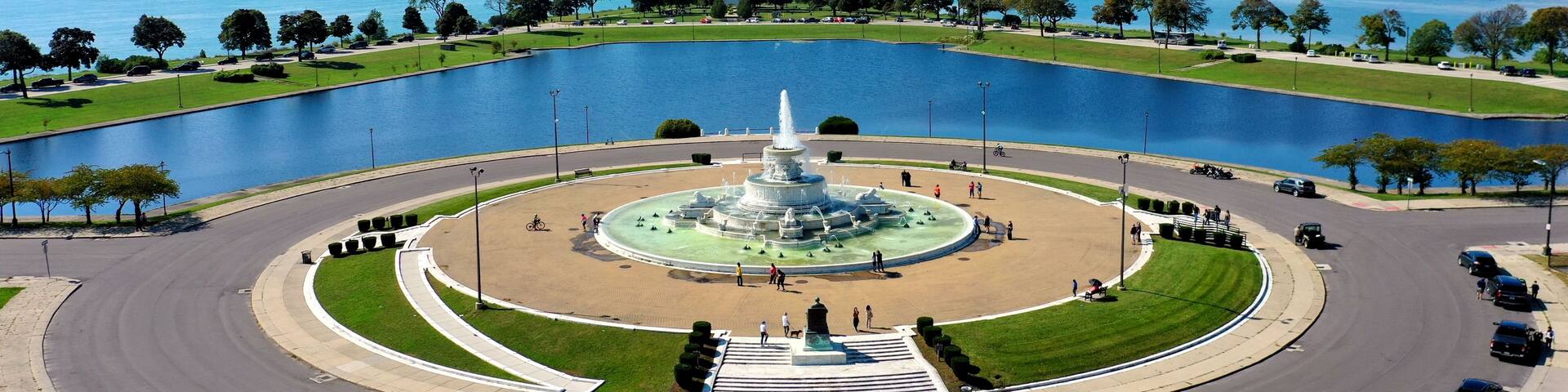 A cityscape with a large body of water, a central fountain, and a bridge in the distance.