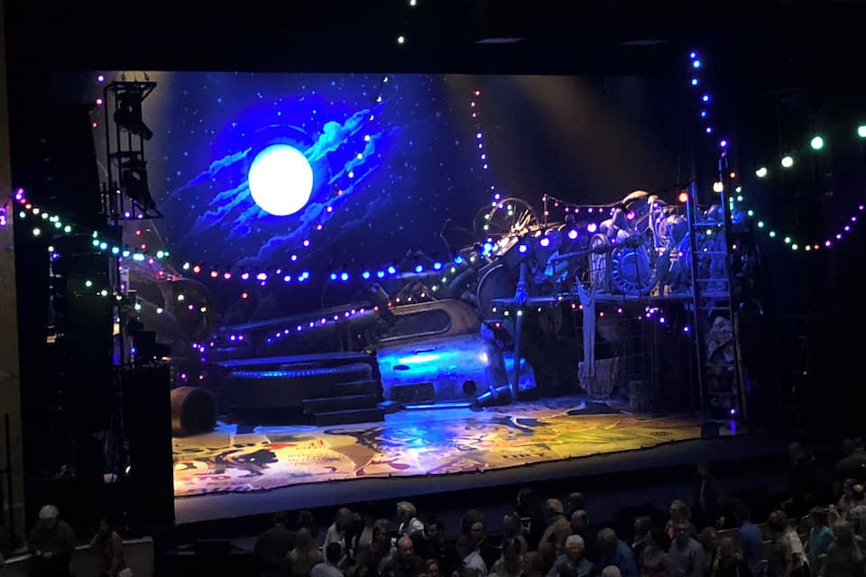 A theater stage with a large screen displaying a space-themed scene, including a bright moon and stars, surrounded by colorful lights.