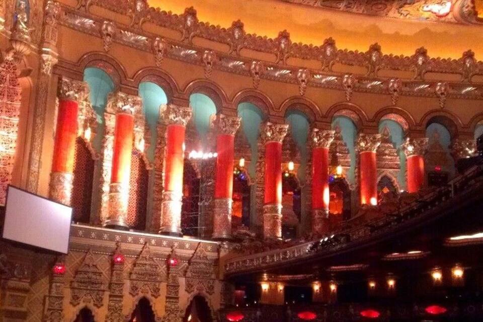 An ornate theater interior with red and teal columns, intricate ceiling designs, and a large screen.