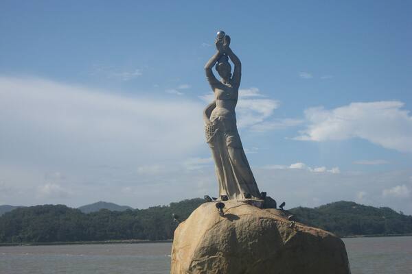 Eine Statue einer Frau auf einem Felsen an einem Gewässer, im Hintergrund Berge.