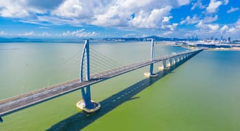 Eine lange Hängebrücke über ruhigem Wasser, im Hintergrund die Skyline einer Stadt.