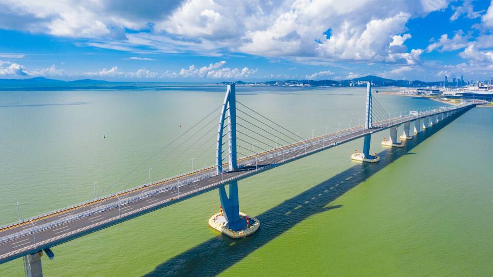 A long suspension bridge over a calm body of water with a city skyline in the background.