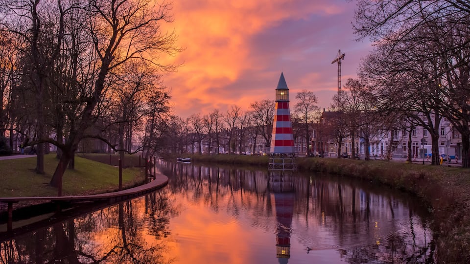 A calm river with a red and white striped lighthouse, surrounded by leafless trees and a sunset sky.