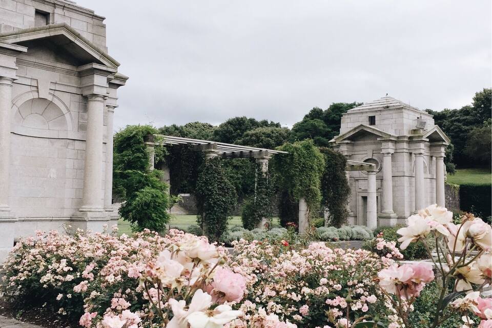 En klassisk byggnad med kolonner och en pergola som är täckt av murgröna, omgiven av frodiga rosa och vita rosor.