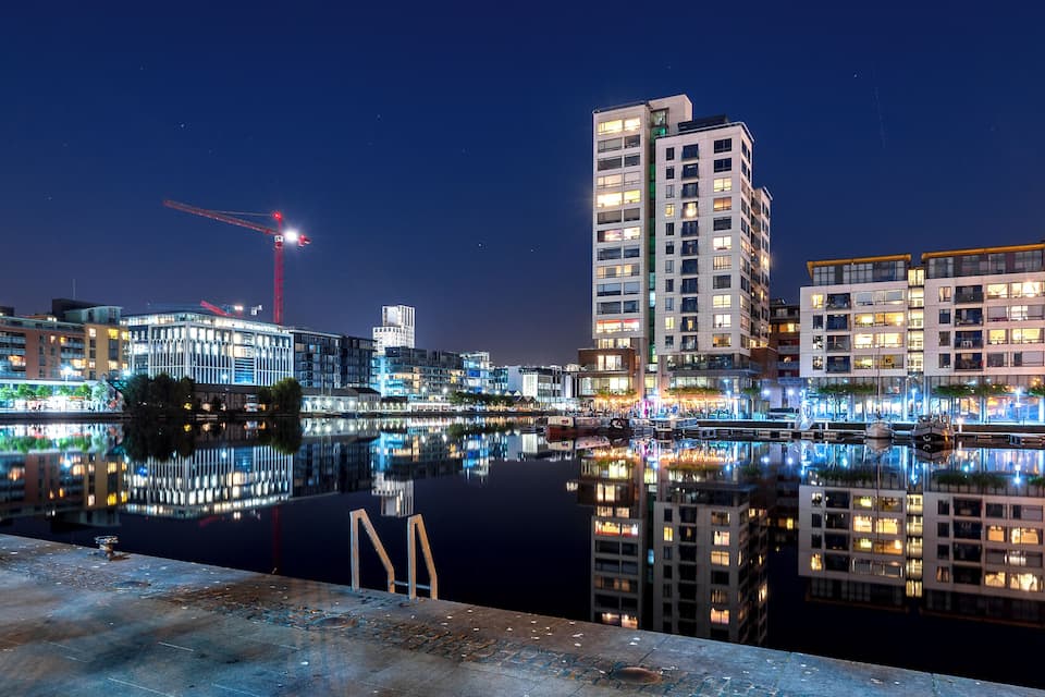 A nighttime cityscape with illuminated buildings reflecting on a calm body of water.