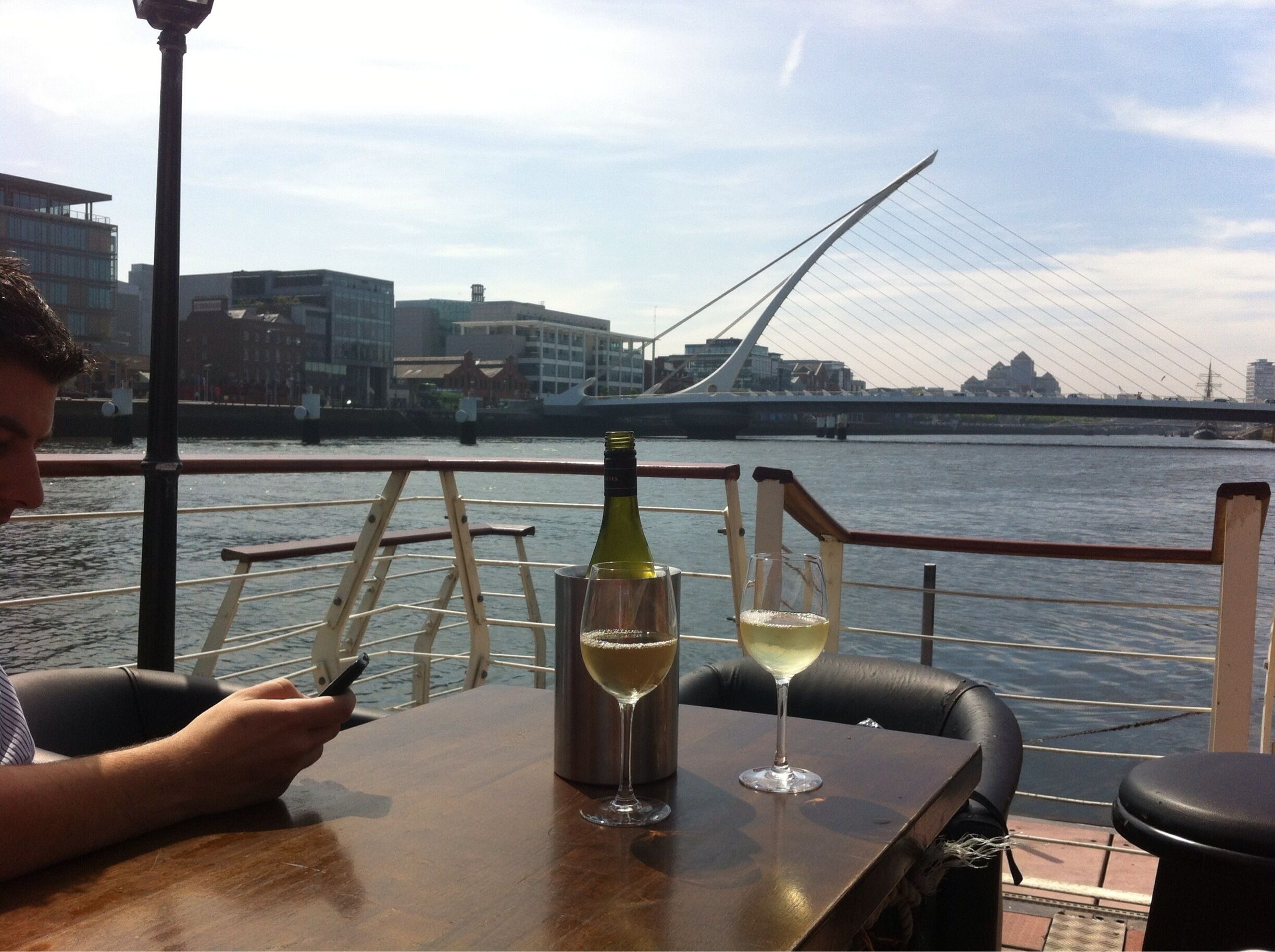 A person sitting at a table by a waterfront with a bottle of wine and a glass of white wine, overlooking a modern bridge and buildings.