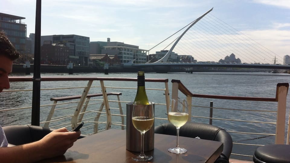 A person sitting at a table by a waterfront with a bottle of wine and a glass of white wine, overlooking a modern bridge and buildings.
