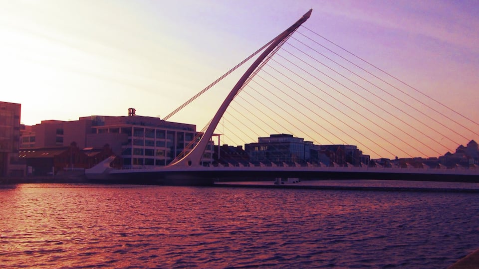 A cable-stayed bridge at sunset with buildings in the background.