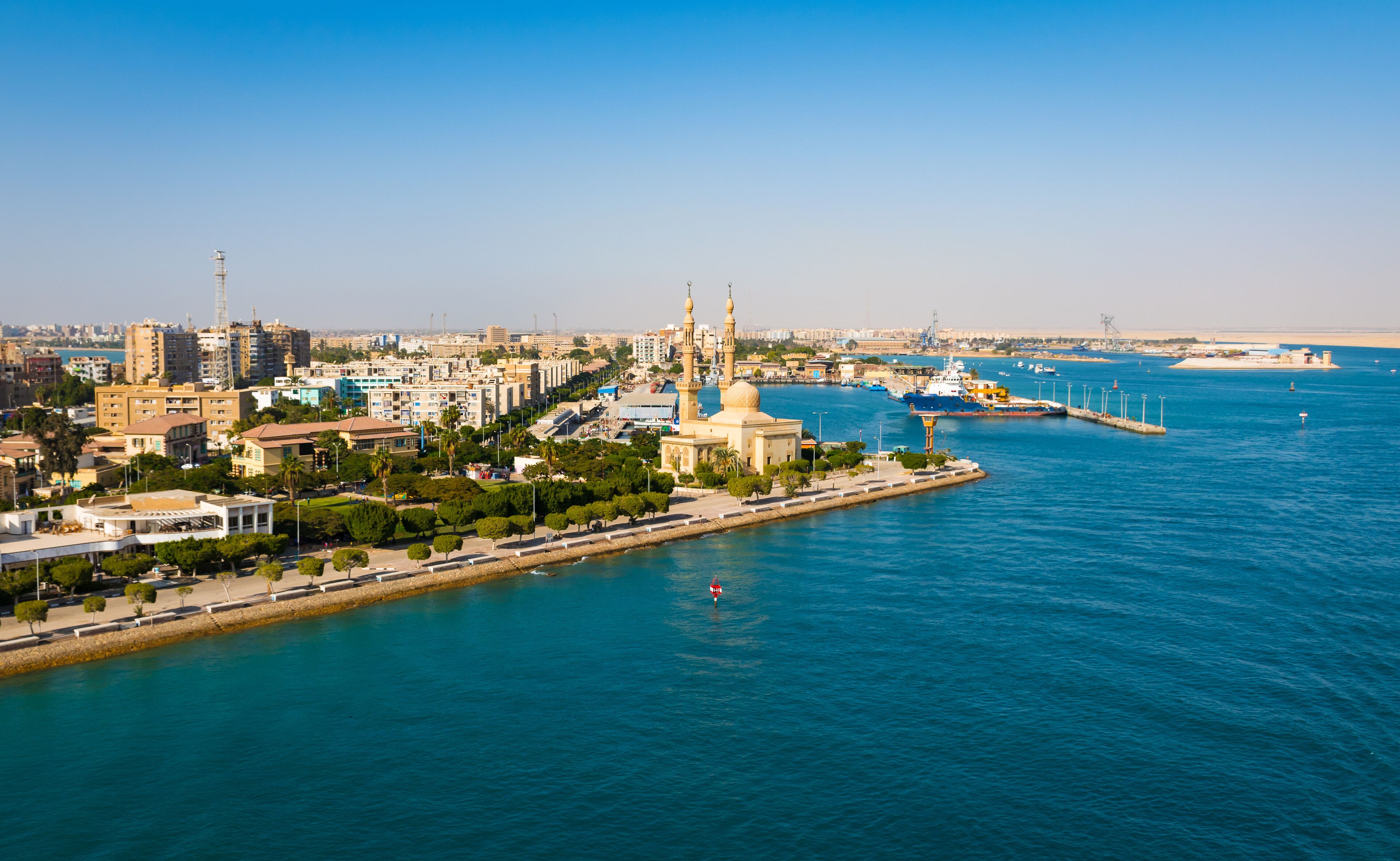 A coastal cityscape with a prominent mosque, a harbor with ships, and a clear blue sky.