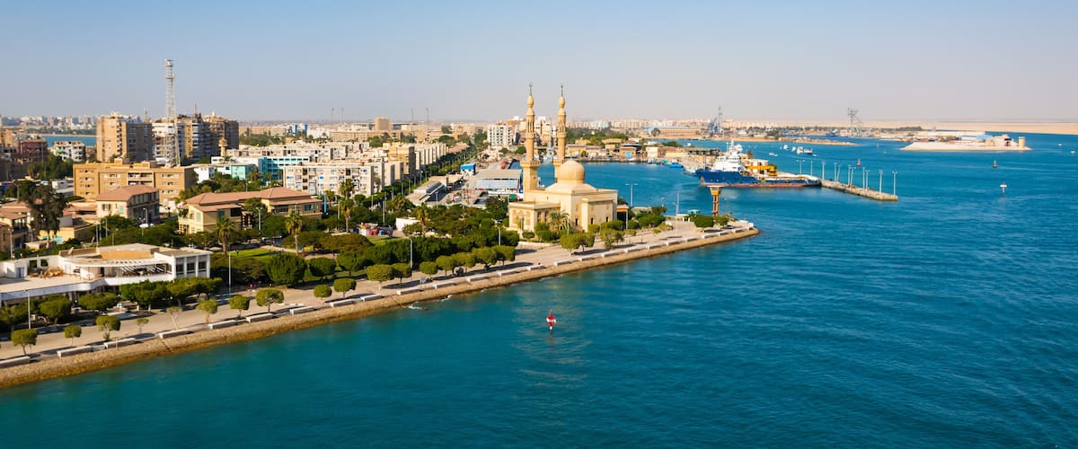 A coastal cityscape with a prominent mosque, a harbor with ships, and a clear blue sky.