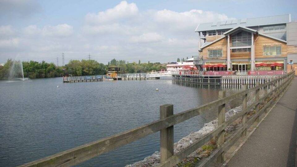 A riverside walkway with a wooden fence, a bridge, and a building with a restaurant.