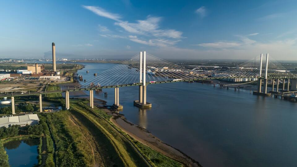 A large cable-stayed bridge spanning a wide river, with industrial buildings and a green area with a pond on the left.
