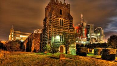 A historic building with a tower, surrounded by modern industrial structures and a grassy area with a well-maintained hedge.