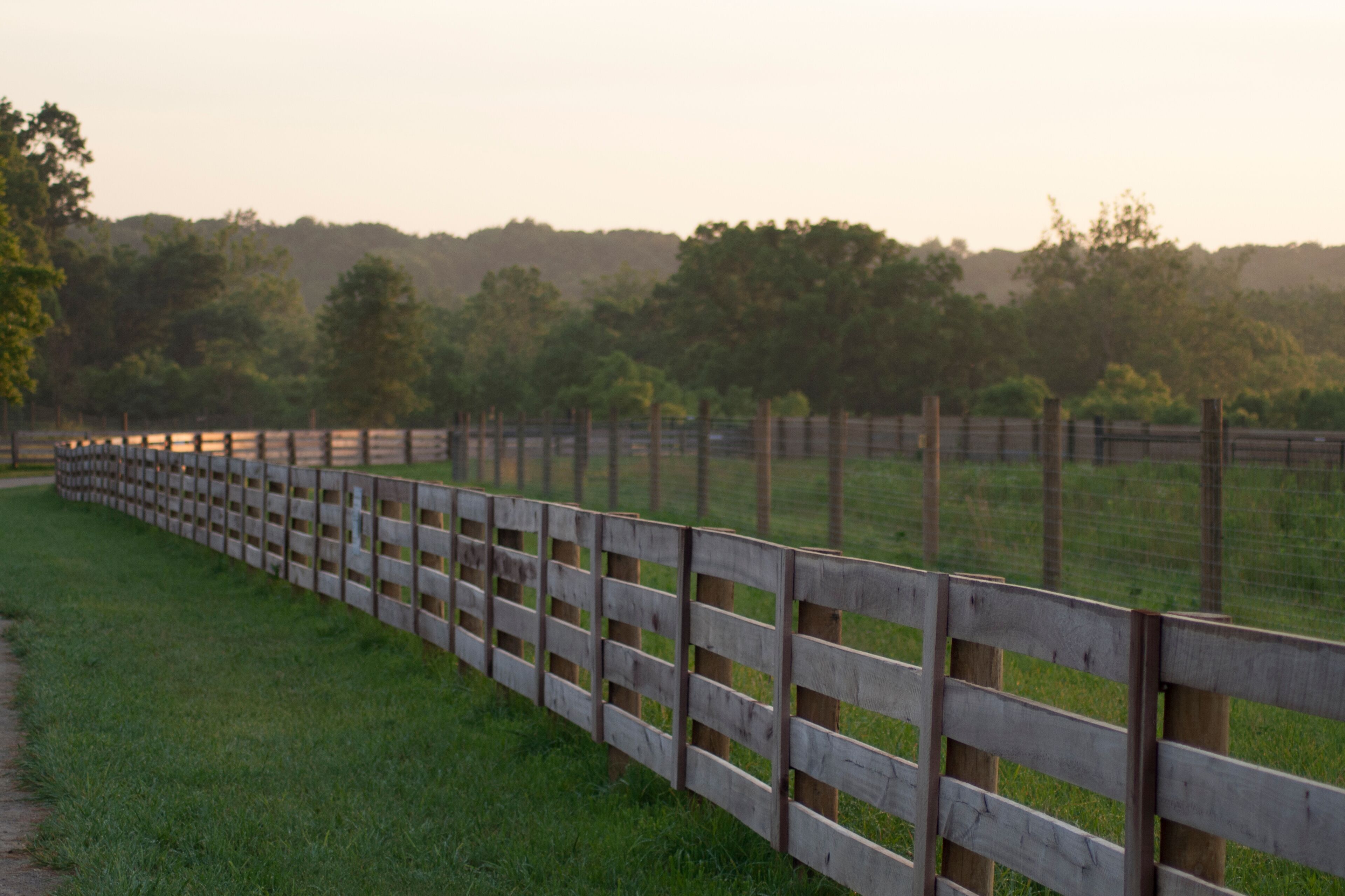 A wooden fence along a grassy path with trees and hills in the background.
