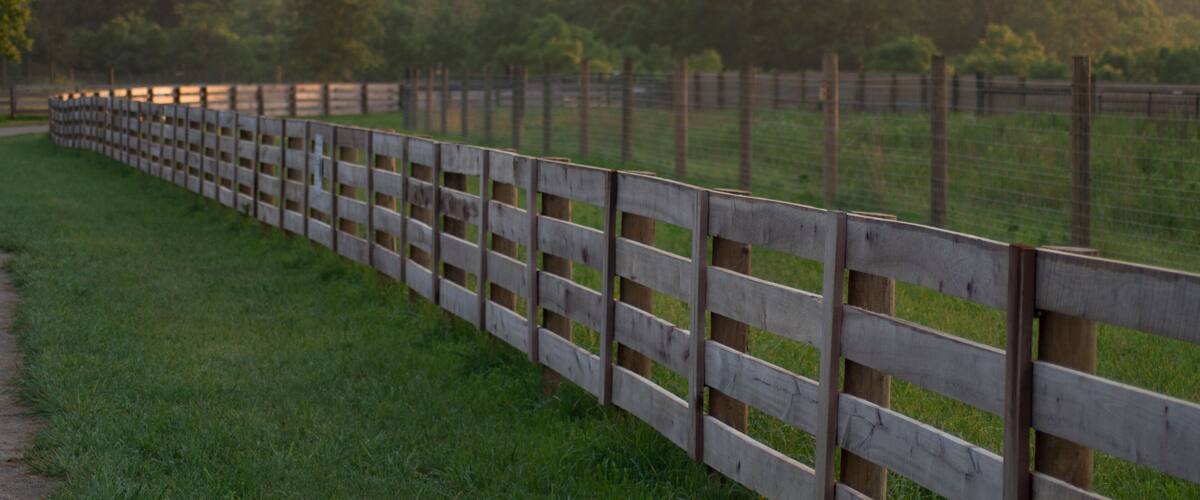 A wooden fence along a grassy path with trees and hills in the background.