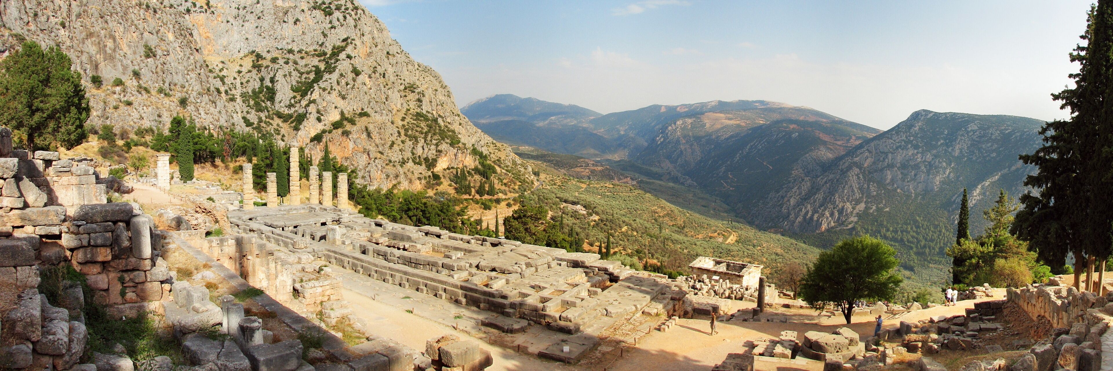 Ancient ruins with columns and stone structures, set against a backdrop of mountains and trees.