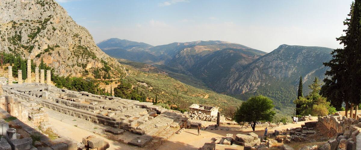 Ancient ruins with columns and stone structures, set against a backdrop of mountains and trees.